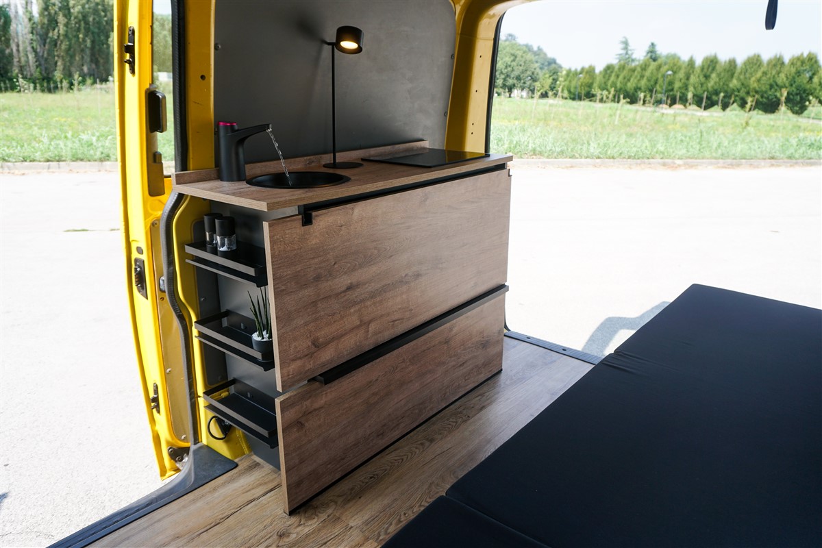 Interno di un Volkswagen T5 giallo ex Deutsche Post camperizzato, con letto aperto 120x190 e mobile cucina in legno e antracite